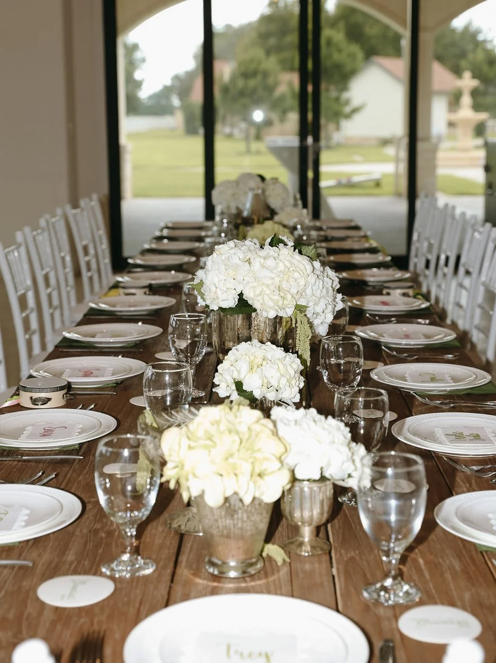 Long dining table decorated with white hydrangea flowers in vintage vases, set with plates, glasses, and utensils, in a room with large windows overlooking a green outdoor landscape.