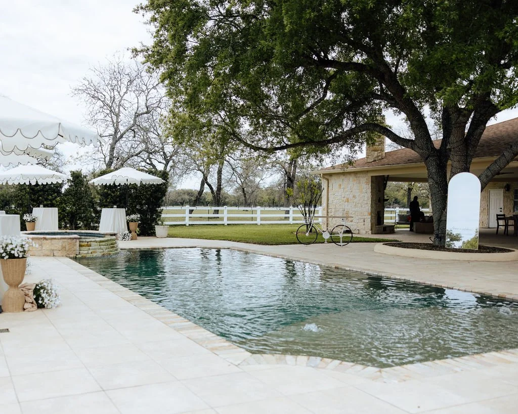 An intimate cocktail hour setting with a swimming pool, white umbrellas, potted plants, green grass, trees, a stone house, a bicycle, and a person in the distance under a tree.
