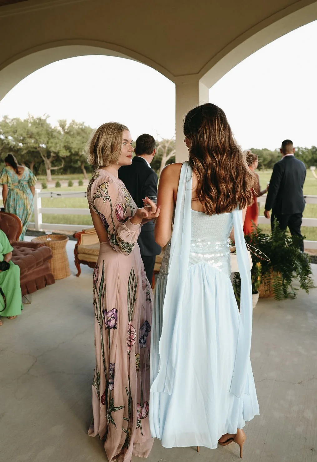 Two women are talking on a porch during a wedding or formal event. One woman is wearing a pink floral dress, and the other is in a white dress. Several other people are in the background, some facing away, dressed in formal attire, with a view of gre