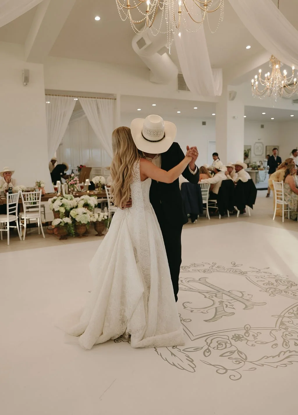 A bride and groom dancing at their wedding reception in the manor with white drapes, chandeliers, and floral arrangements.