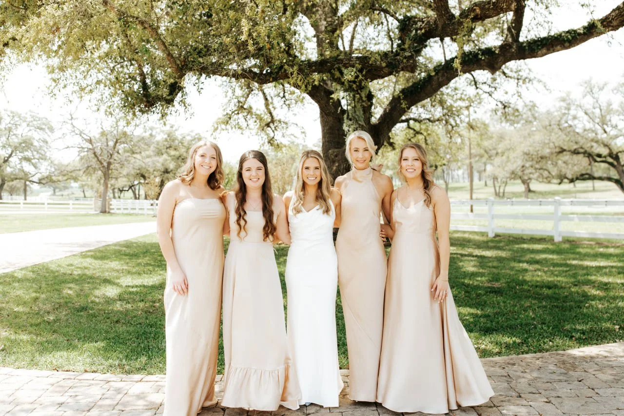 Four bridesmaids and a bride are standing outdoors under a large oak tree, smiling together at a wedding.