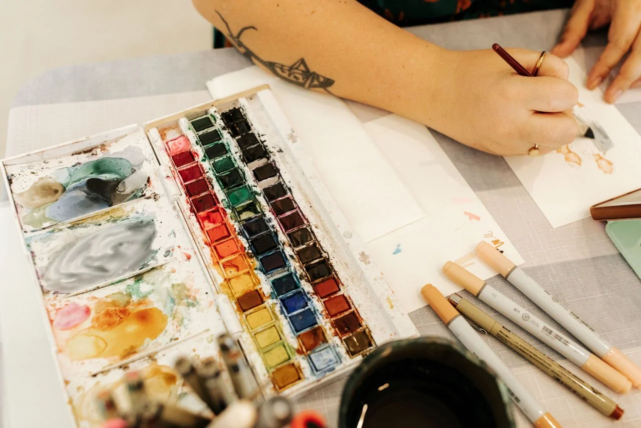 Artist painting guest portraits at Ro Manor during a wedding cocktail hour. Hands painting with watercolor paints on paper, with watercolor palette, brushes, and a cup of coffee on the table.
