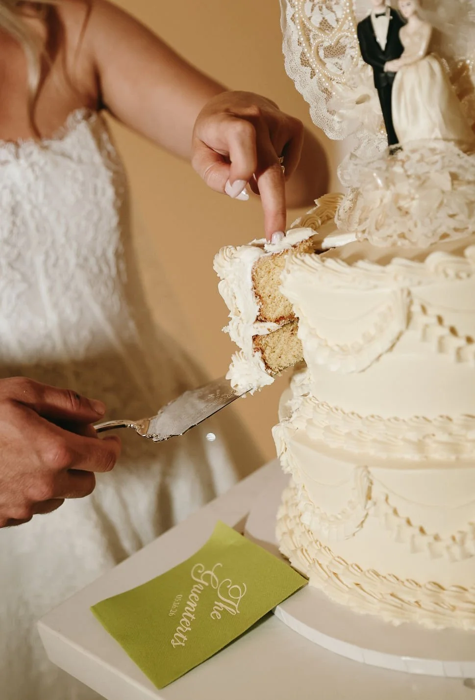 Wedding cake being cut by a bride, with wedding figurines on top, and a custom cocktail napkin at a wedding reception.