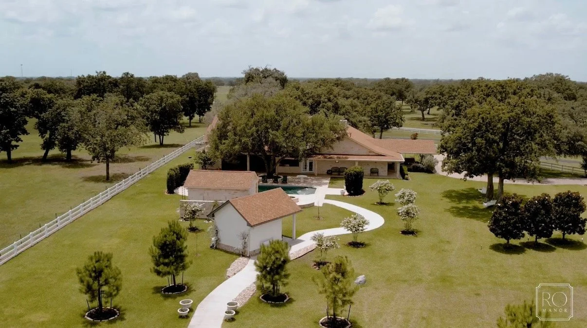 Aerial view of a large rural property with a house, a smaller building, a winding concrete pathway, a fenced area, and many trees on a sunny day.