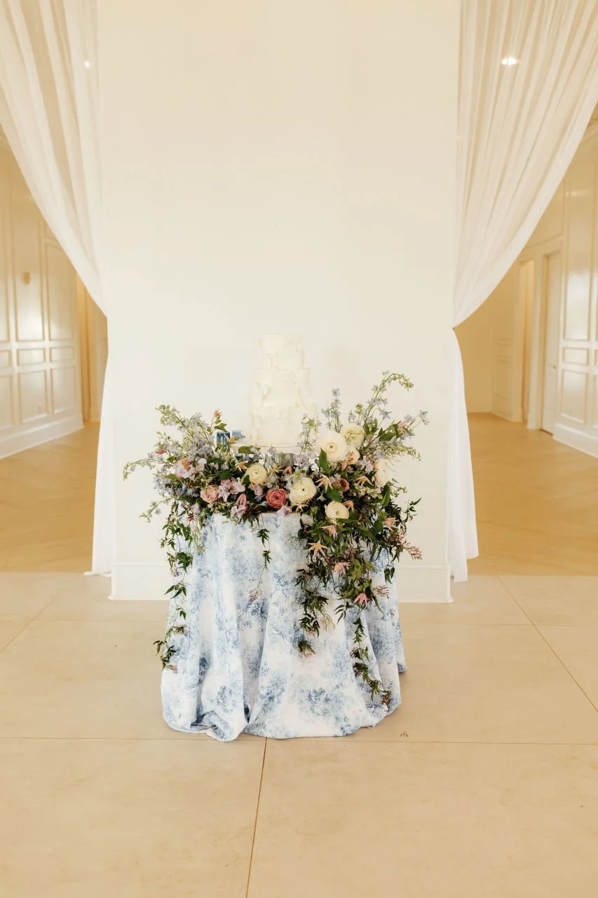A wedding cake with floral decorations on a table covered with a white and blue cloth, set against a light-colored backdrop with curtains.