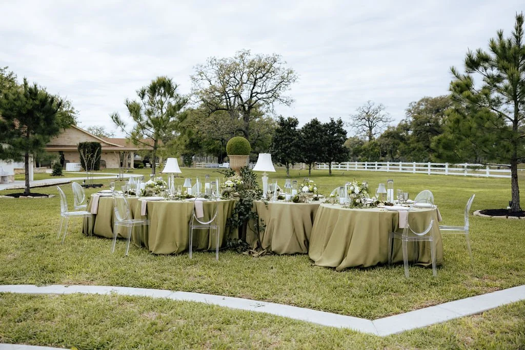 An outdoor banquet caterpillar style table setup on a grassy lawn with a white fence and trees in the background, featuring olive green tablecloths, floral centerpieces, and white lamps.