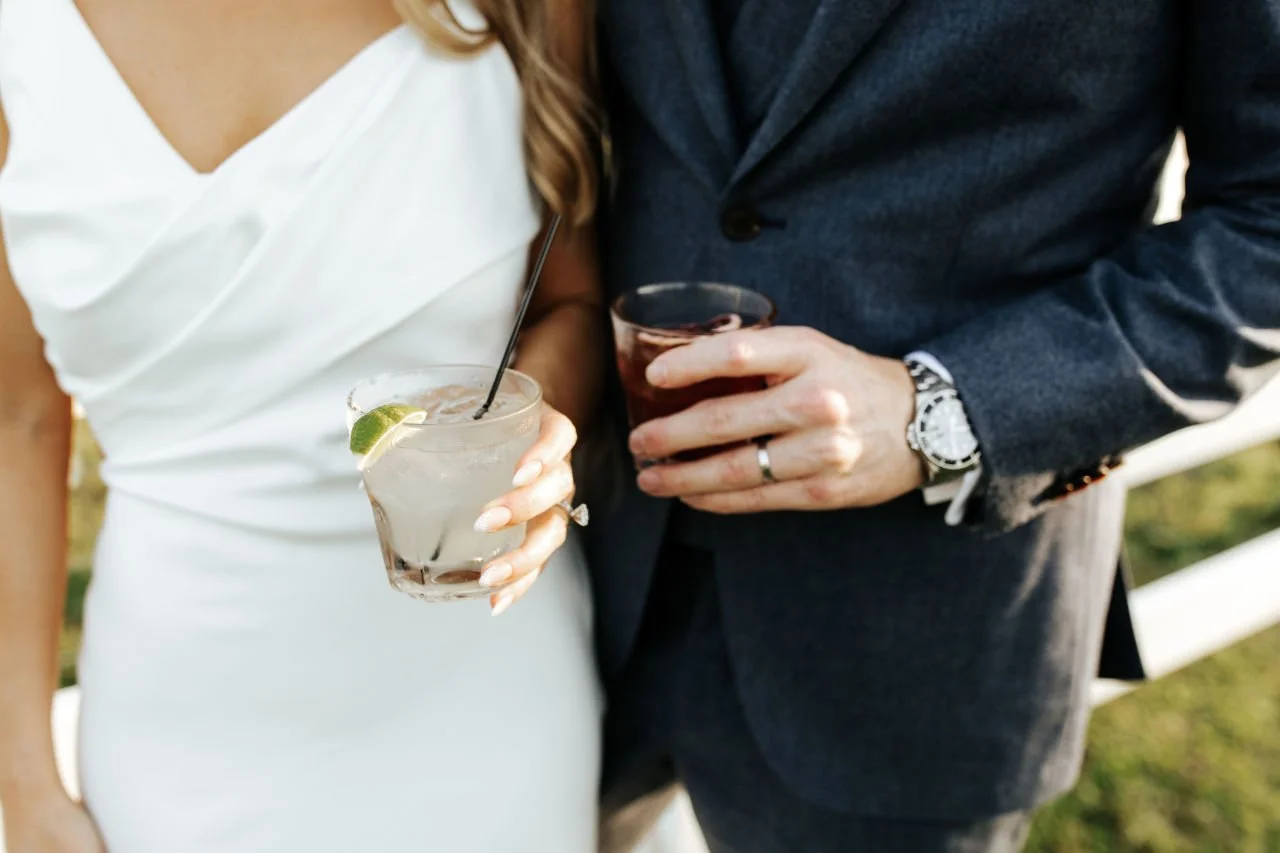 A bride and groom holding cocktails, with the woman in a white dress and the man in a dark suit, outdoors during daytime.