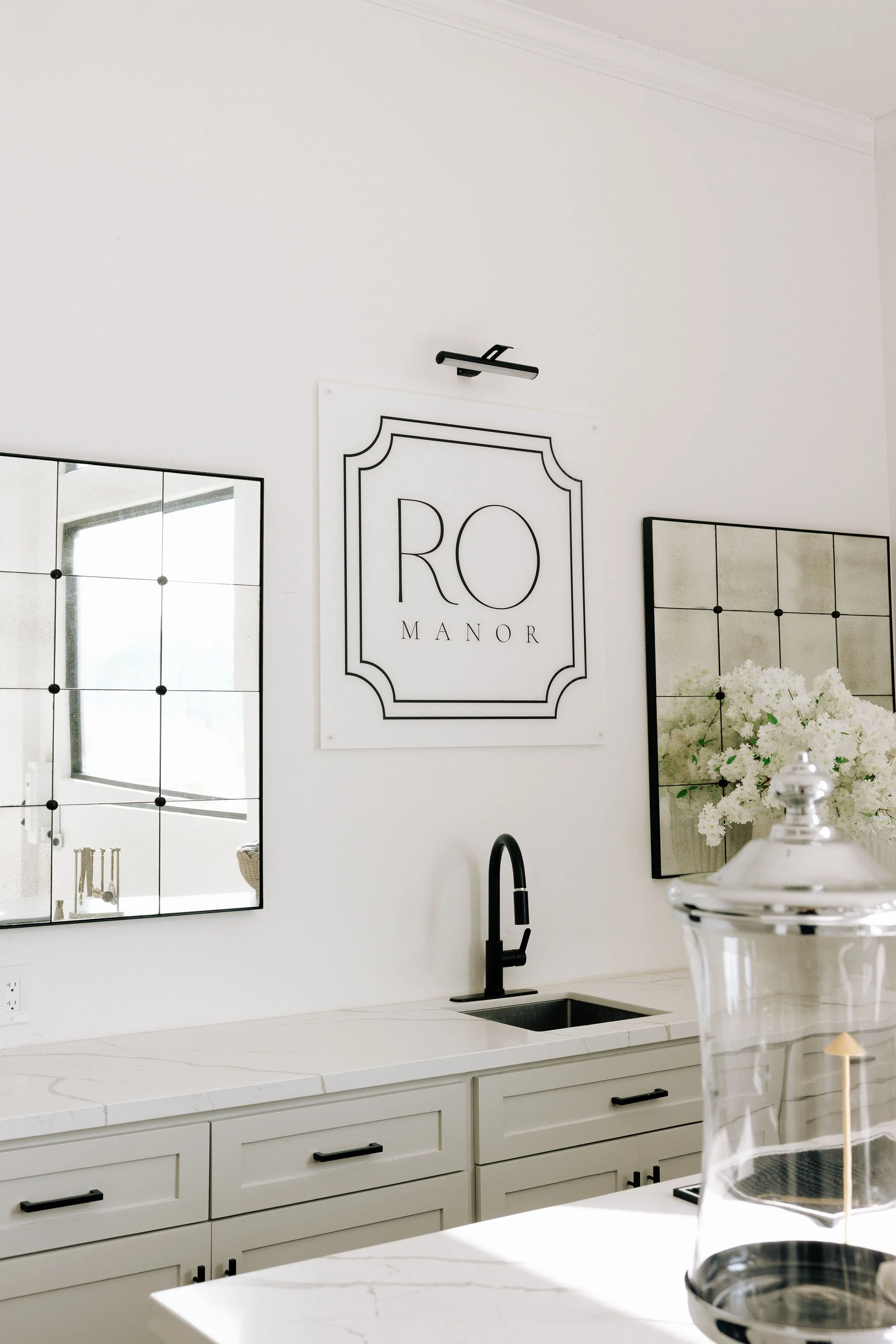 A modern venue bar with white cabinetry, a black faucet, black cabinet handles, and decorative wall mirrors, featuring a sign that says 'RO MANOR' and a bouquet of white flowers on the counter.