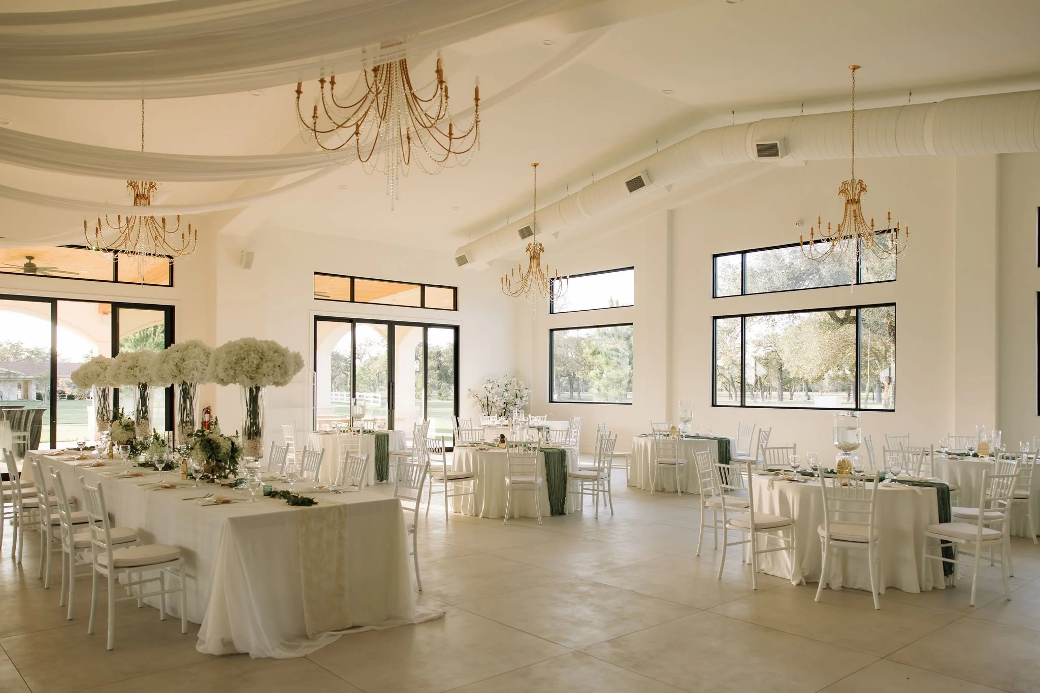 Elegant banquet hall decorated for a wedding reception with white tablecloths, white chairs, floral centerpieces, chandeliers, and large windows letting in natural light.