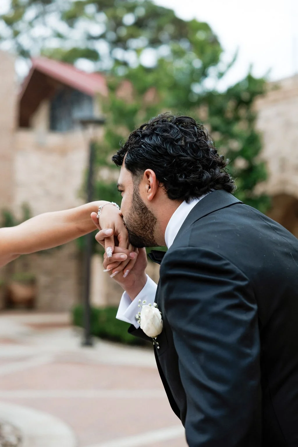 Groom dressed in a tuxedo kisses his wife's hand, outdoor setting with trees and buildings in the background.
