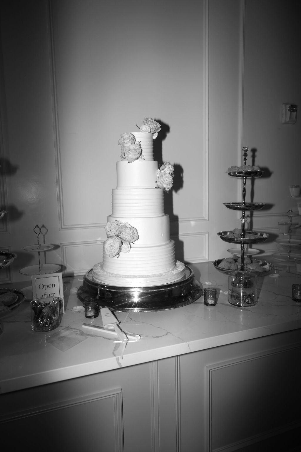 A tall, multi-tiered wedding cake decorated with flowers on a table, flanked by serving platters and a small sign' in a decorated room.
