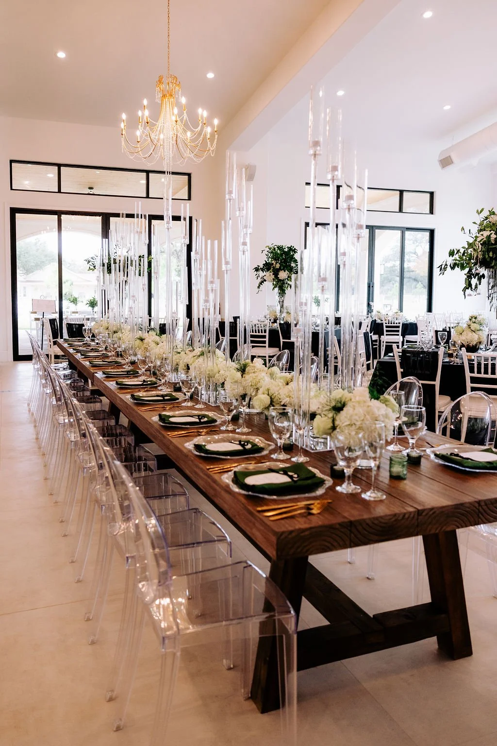 Elegant banquet hall with a long wooden table decorated with white flowers, candles, and greenery, set with black and white plates, glassware, and gold cutlery, surrounded by transparent chairs, illuminated by a chandelier and large windows.