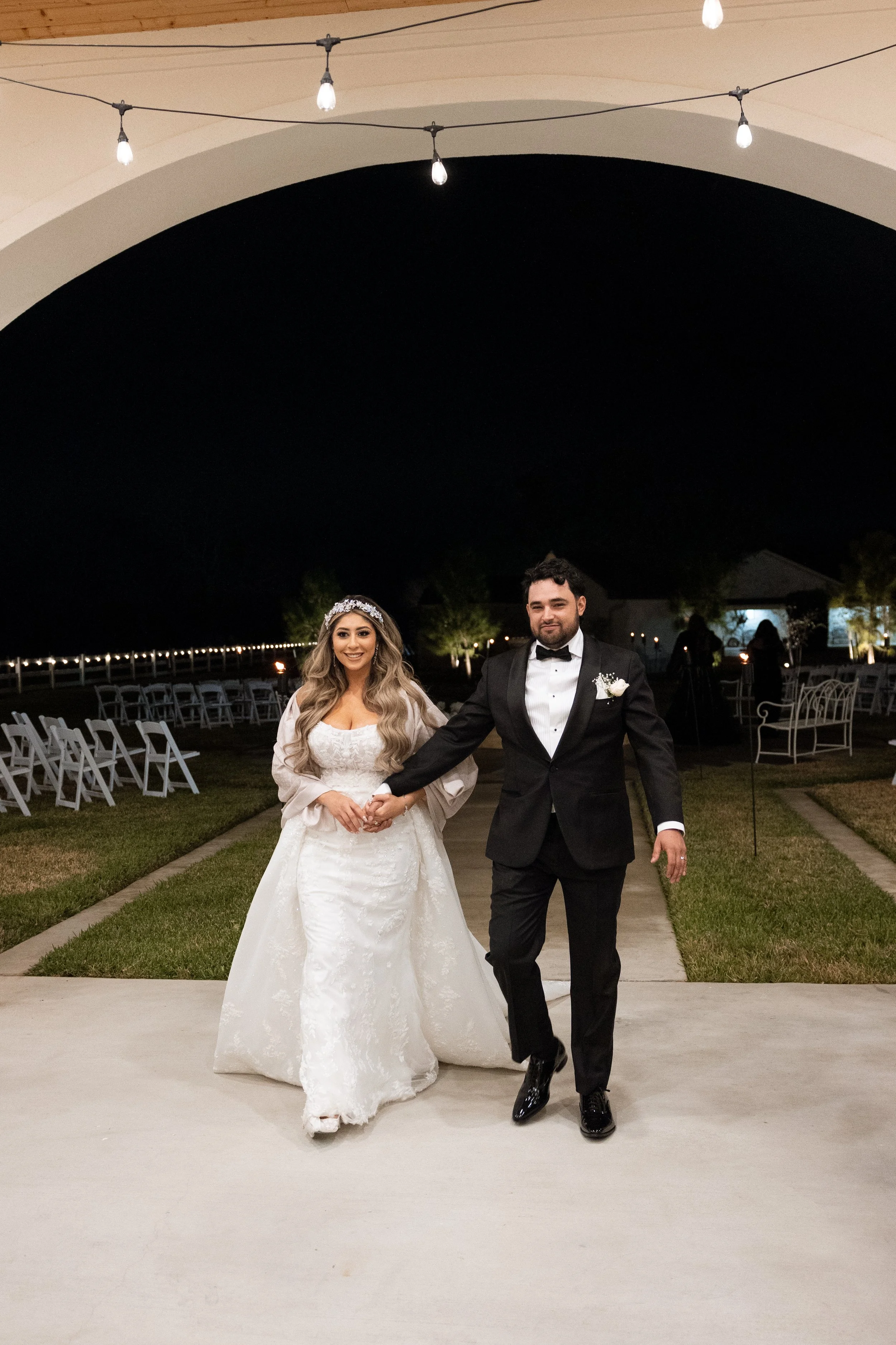 A bride and groom holding hands and dancing under a large white arch decorated with string lights at an outdoor wedding reception at night. The bride is wearing a white lace strapless wedding gown and a floral headpiece, while the groom is dressed in