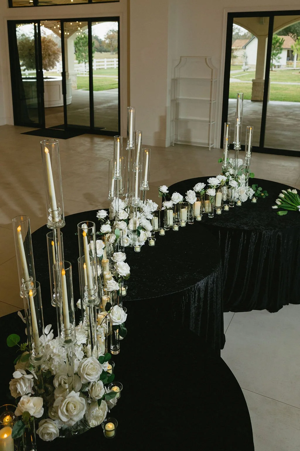 Elegant curved black table with white floral arrangements, tall glass candle holders with lit candles, and small votive candles inside glass holders and roses in a bright indoor space with sliding glass doors opening to a green outdoor area.
