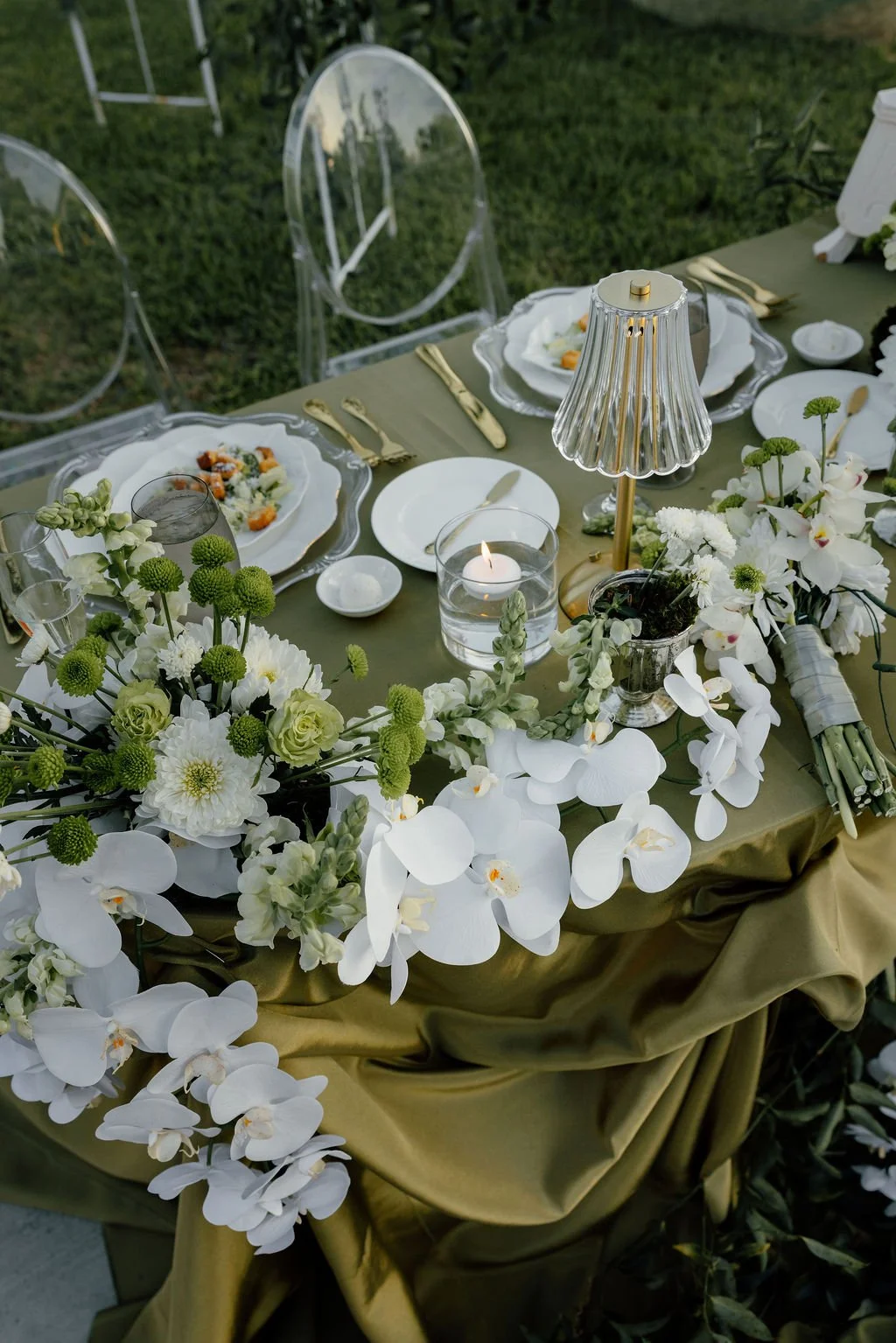 Elegant outdoor dining table decorated with white flowers, gold utensils, a glass lamp, a candle, and place settings with plates and glasses, set on an olive tablecloth in a garden wedding.