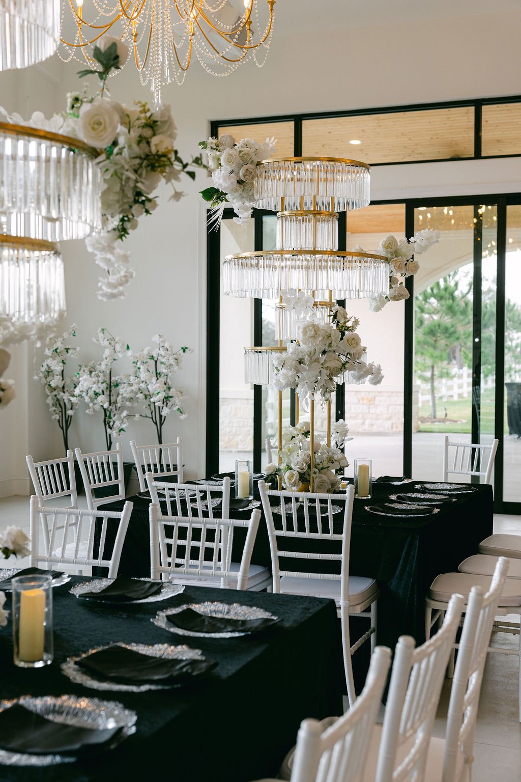 Elegant dining setup with black tablecloths, white chairs, and tall floral centerpieces adorned with white roses and orchids under large crystal chandeliers.