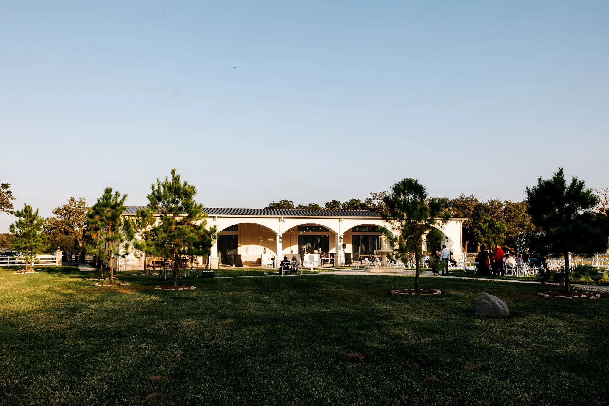 An outdoor event space with a building featuring arched openings, surrounded by green grass and trees, with people gathered and chairs arranged for an event.