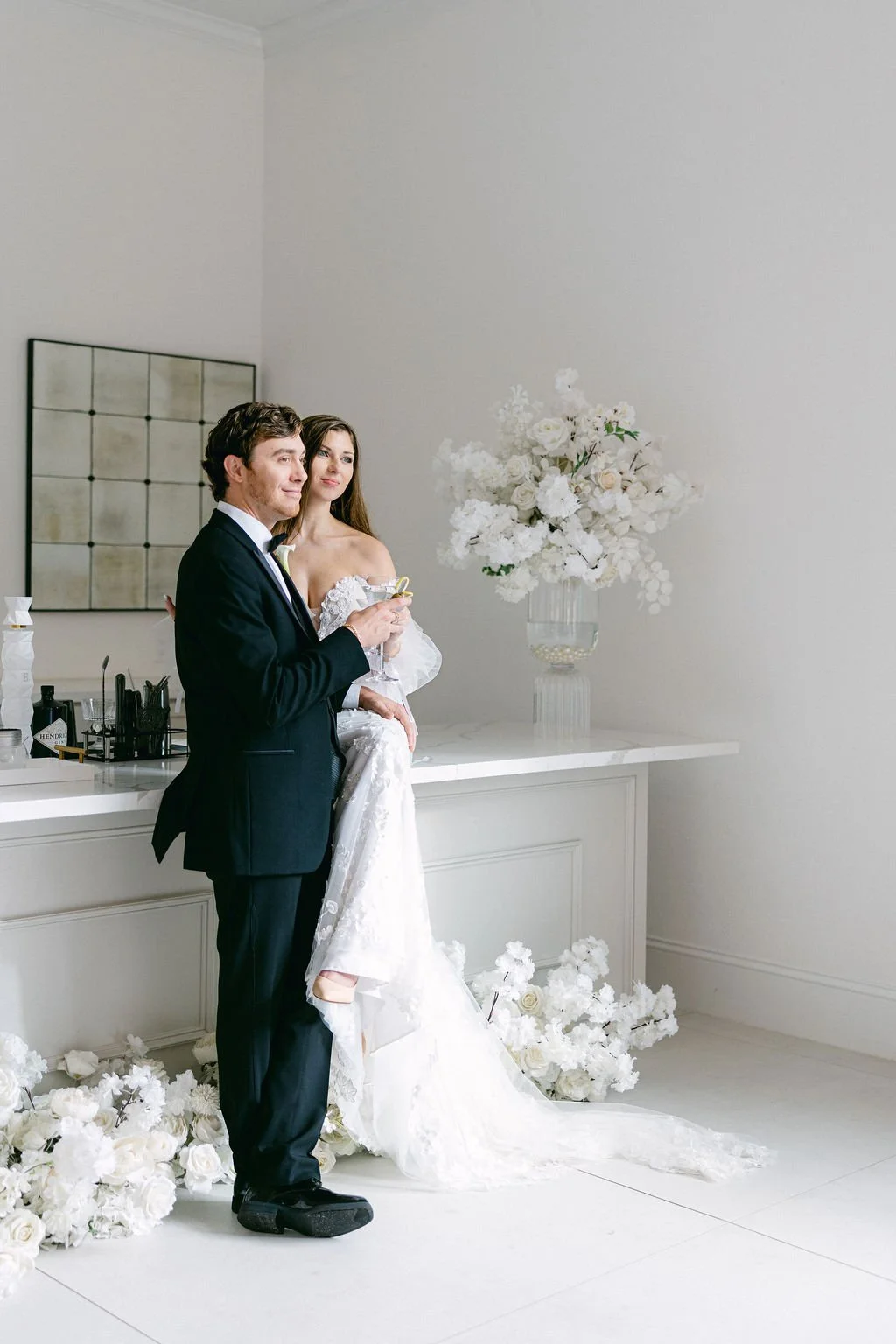 A newlywed couple in wedding attire standing in a room with white walls and decor. The groom is holding a drink, and the bride is sitting on his lap. There are white flowers and a large floral arrangement in a glass vase on the counter behind them.