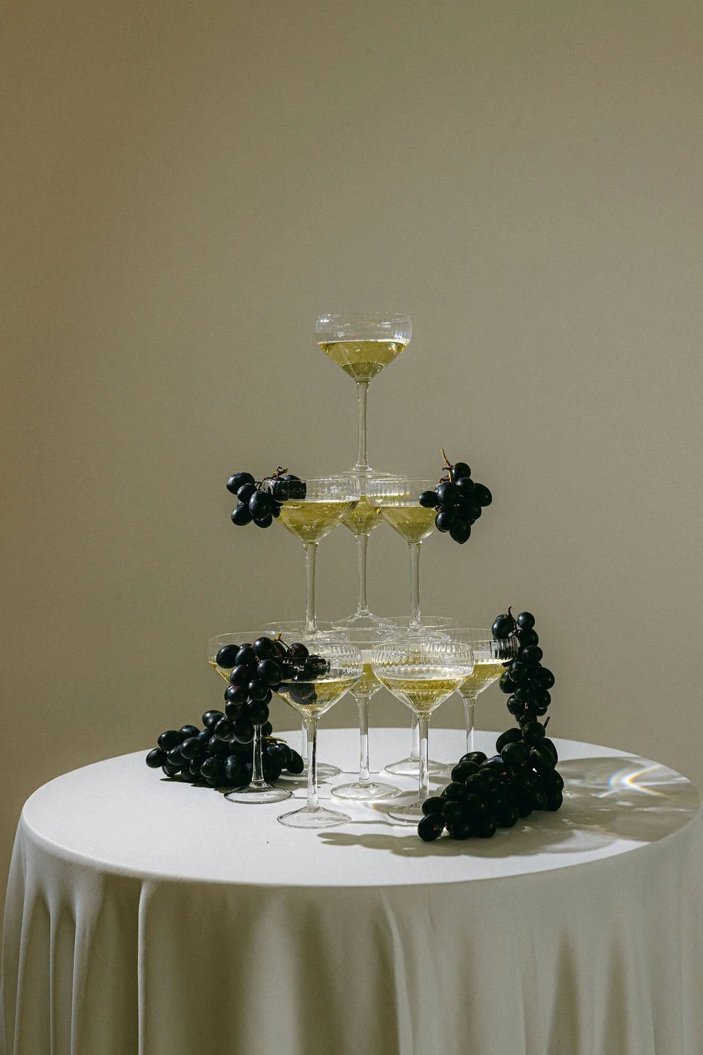 A pyramid of seven champagne glasses filled with champagne, decorated with black grapes, on a white table with a plain background.