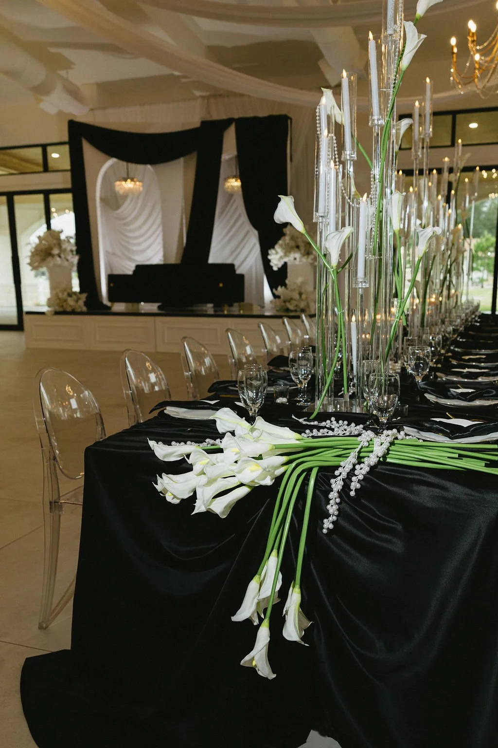 Elegant event table with black tablecloth decorated with white calla lilies and tall glass candleholders, set up for a wedding reception or formal dinner in a well-lit banquet hall with floral arrangements.