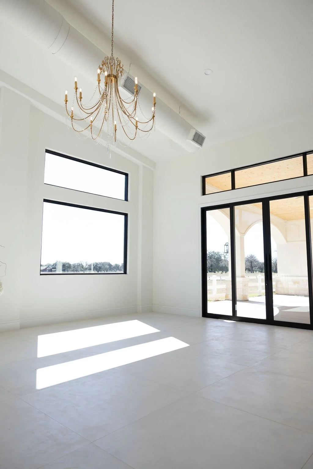 Bright, empty room with large windows, sliding glass door, and a gold chandelier.