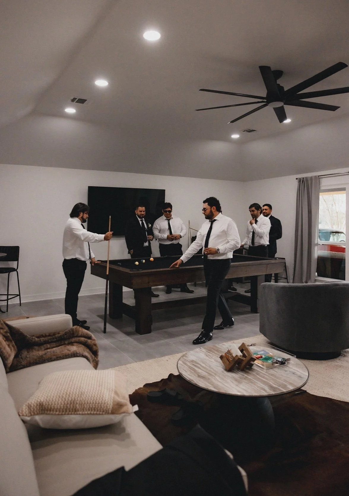 A group of six men in formal attire playing billiards in a modern living room with a pool table, large window with curtains, and a flat-screen TV on the wall.