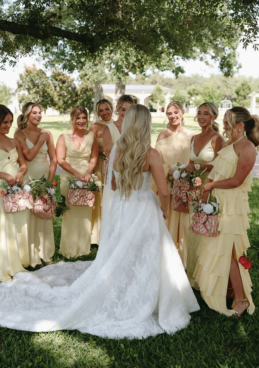 A bride in a white lace wedding dress standing outdoors under a tree, surrounded by her bridesmaids in yellow dresses, all smiling and holding custom floral purse bouquets at a wedding ceremony.