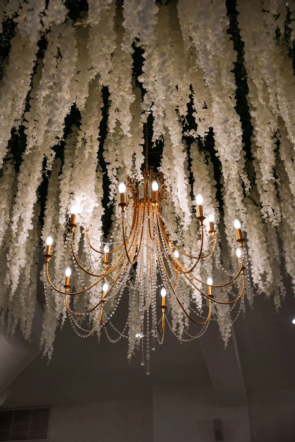 A gold chandelier hanging from a ceiling, surrounded by draped white wedding flowers and hanging crystal beads.