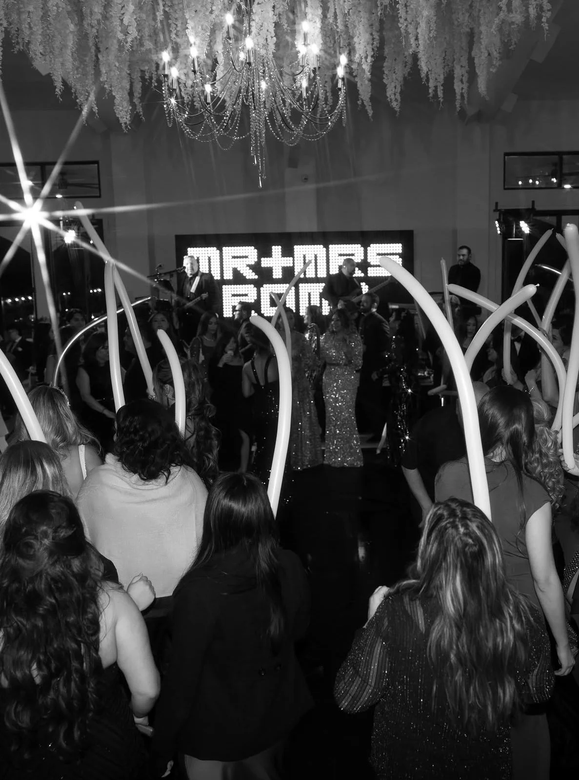 A black and white photo of a wedding celebration with a crowd dancing and a live band performing on stage, illuminated by a large chandelier and decorative hanging flowers.