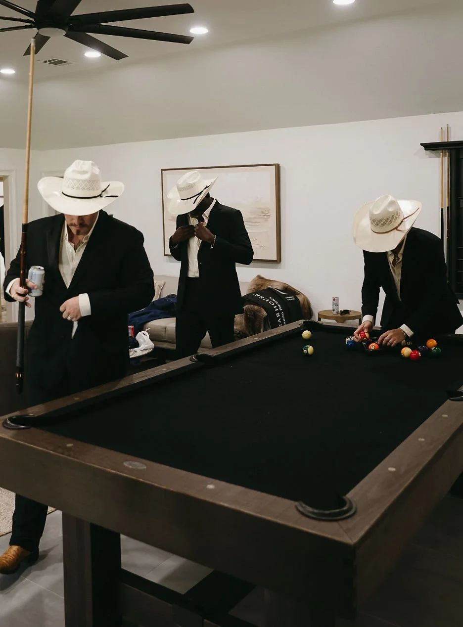 Three men in suits and white hats playing pool in a room with white walls, a ceiling fan, and a sofa in the background.