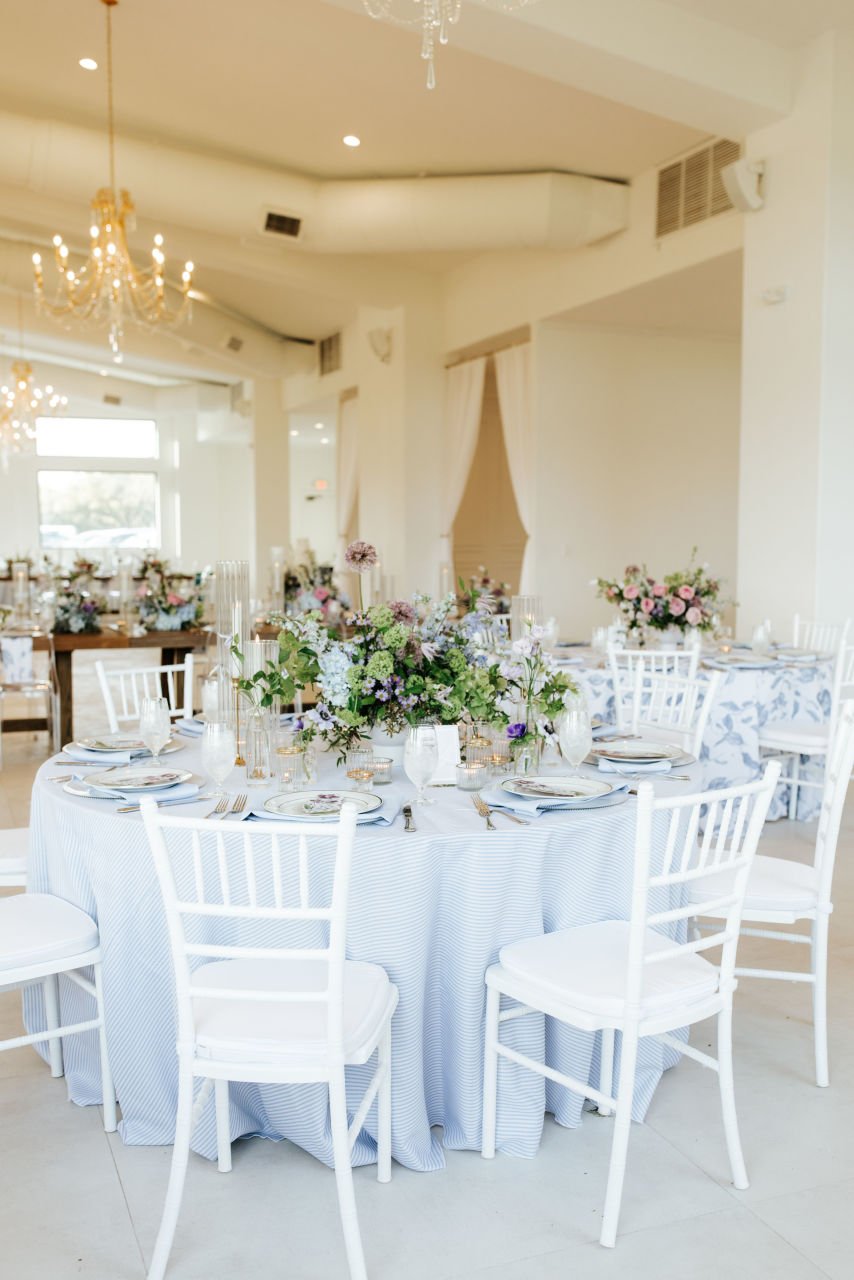 Elegant banquet hall decorated with chandeliers, white tables, and floral centerpieces with pink, purple, blue and white flowers in a soft, light-filled space.