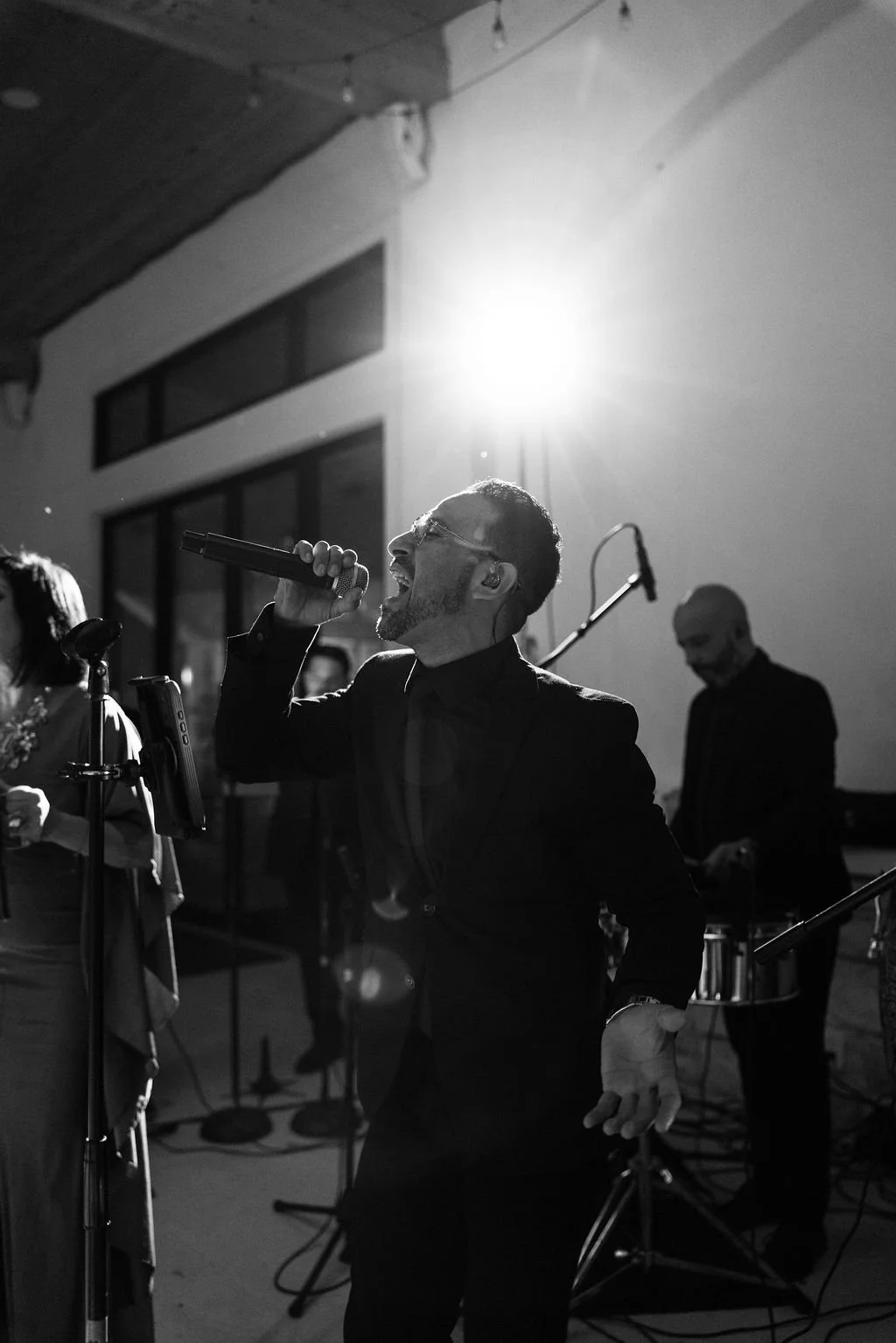 A black and white photo of a man singing passionately into a microphone during a live music performance at an outdoor wedding reception, with a drummer visible in the background.