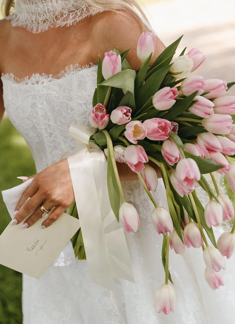 A bride holding a bouquet of pink and white tulips, wearing a white lace wedding dress and a wedding ring, with a note in her hand that says "KALEJ".