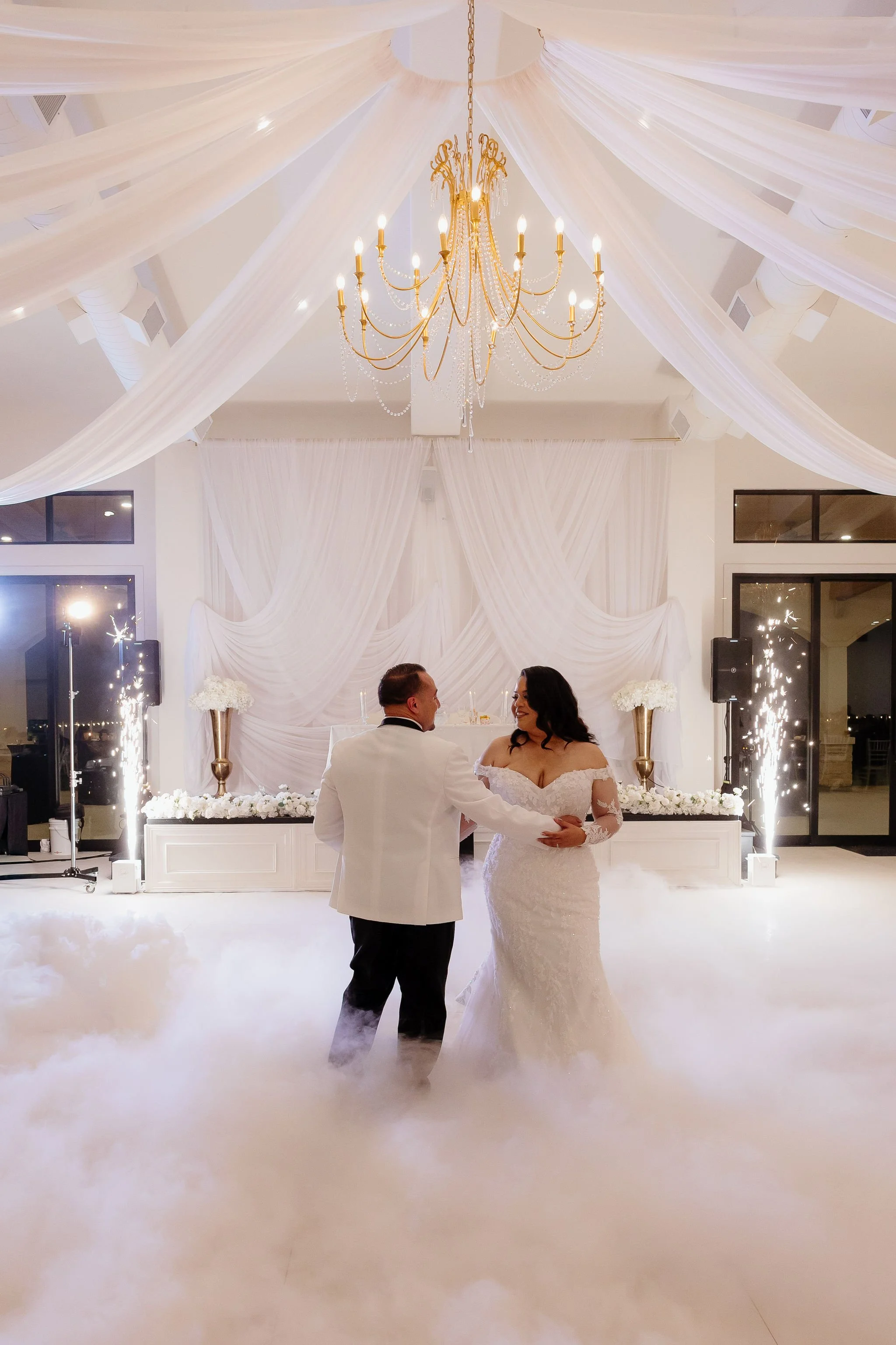 A bride and groom are dancing in a decorated wedding reception hall at Ro Manor with draped white fabric and a large chandelier hanging from the ceiling. There are sparklers and floral arrangements in the background and a dancing on the clouds effect