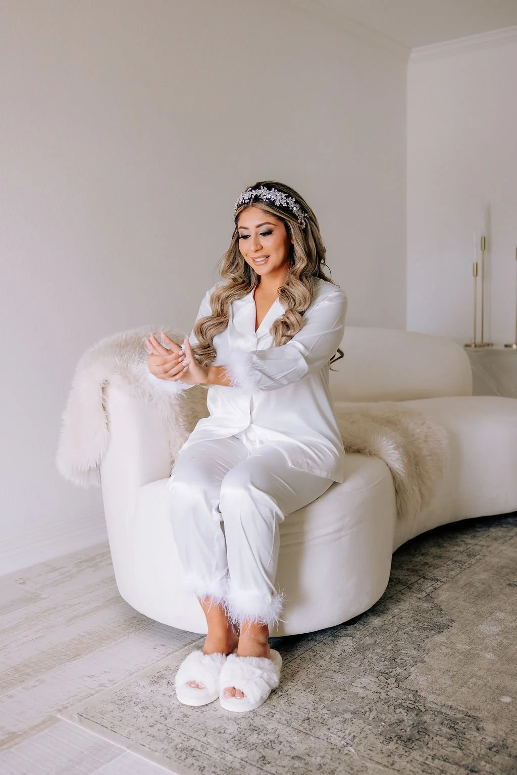 Bride in the bridal suite in white satin pajamas sitting on a modern, white, curved sofa with a cream fur blanket, in a minimalist bedroom. She has long, wavy hair, wearing a silver floral headband, and fluffy slippers.