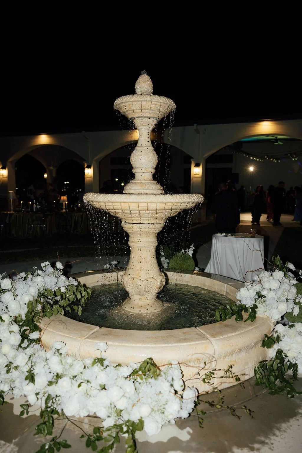 A lit outdoor fountain decorated with white flowers at night, with people dancing and socializing in the background.