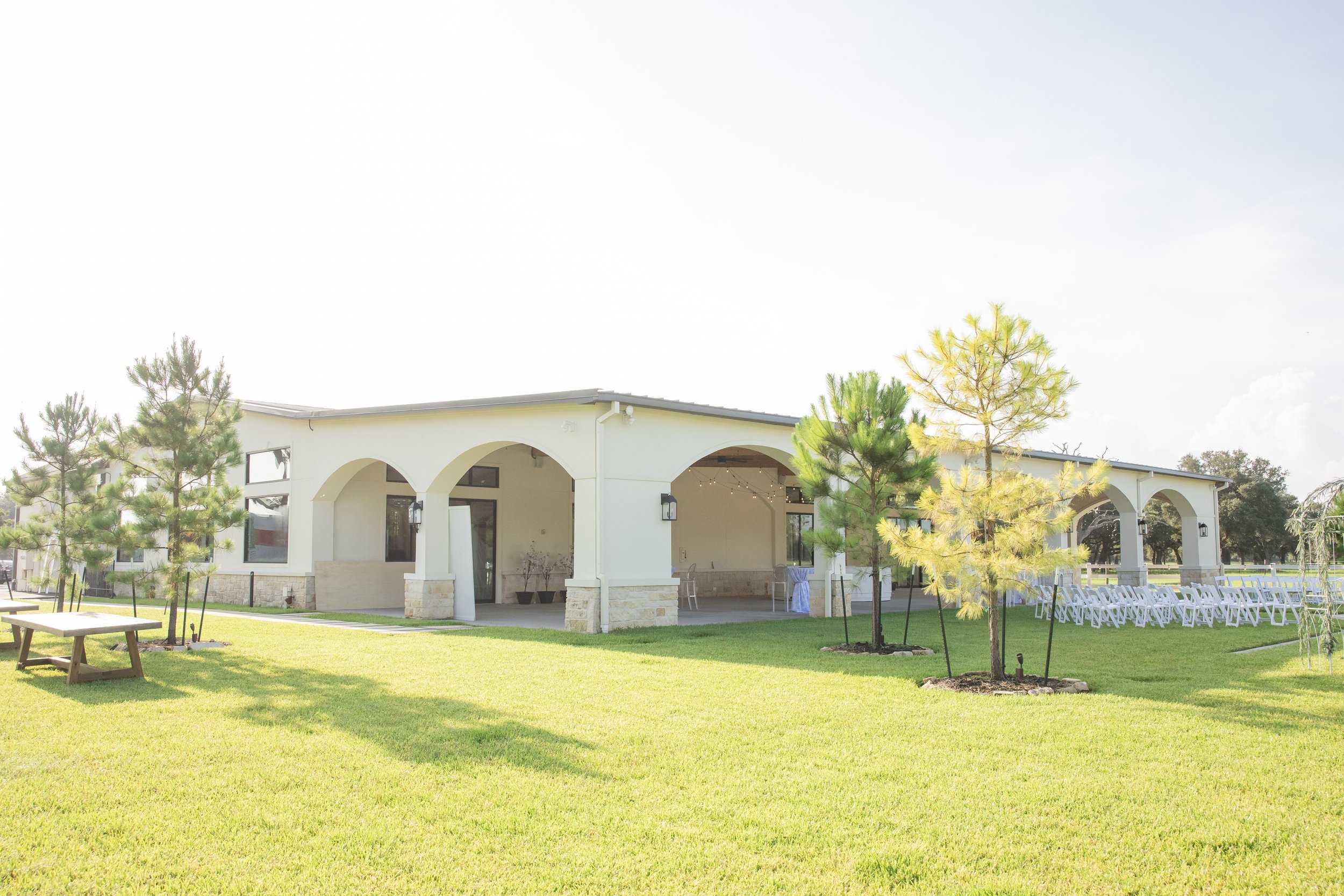 A white house with an arched porch and large windows, surrounded by a well-maintained lawn and several young trees, with white chairs set up on the grass for an outdoor event.