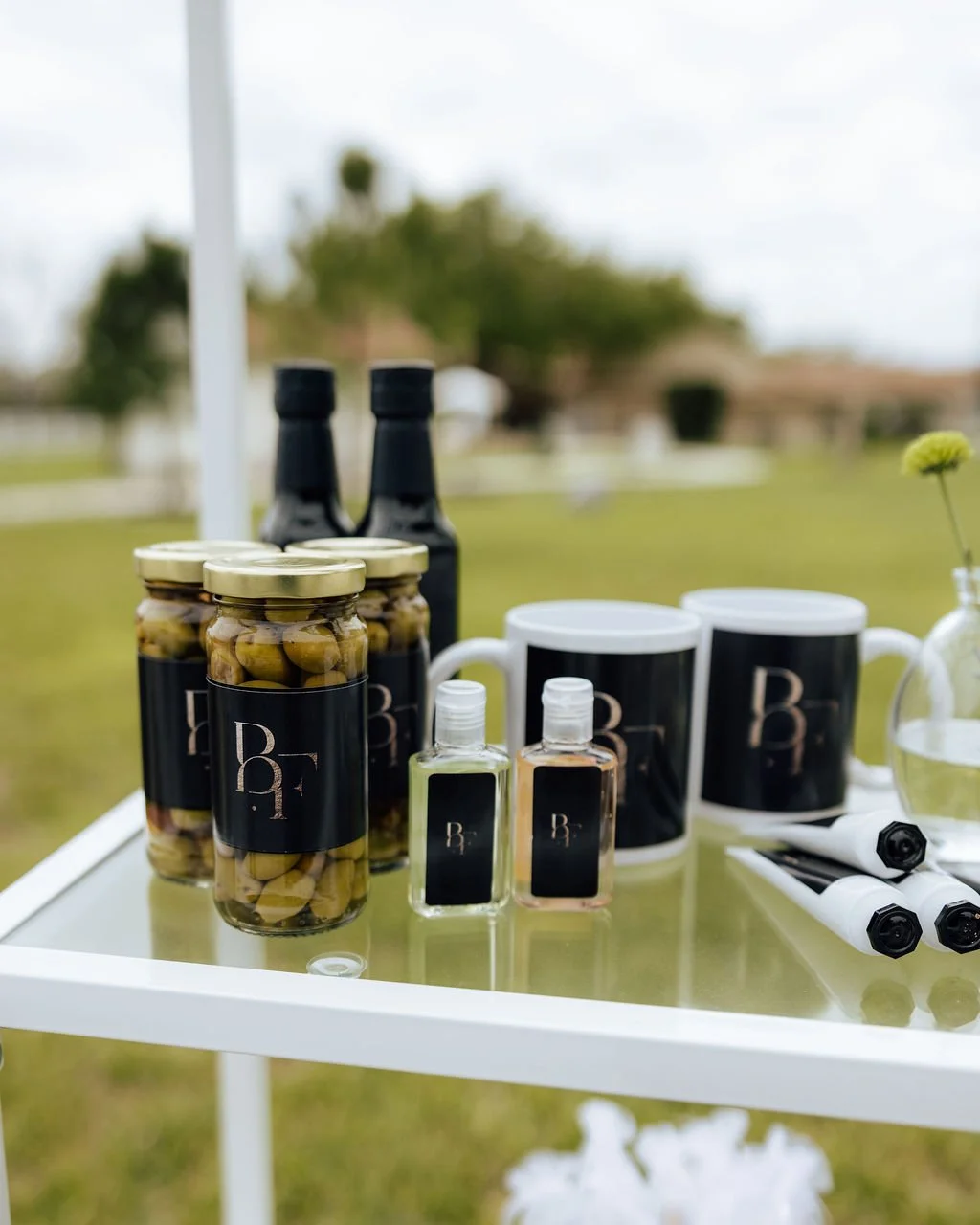 Display of bottled beverages, jars of olives, stainless steel cups, and small bottles arranged on a white table outdoors, with blurred trees and grass in the background. Wedding favor display on shelf.