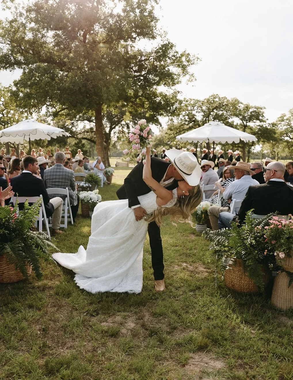 A bride and groom share a kiss during an outdoor wedding ceremony, with the groom dipping the bride and holding a bouquet of pink flowers, surrounded by guests seated under white umbrellas and majestic oak trees.
