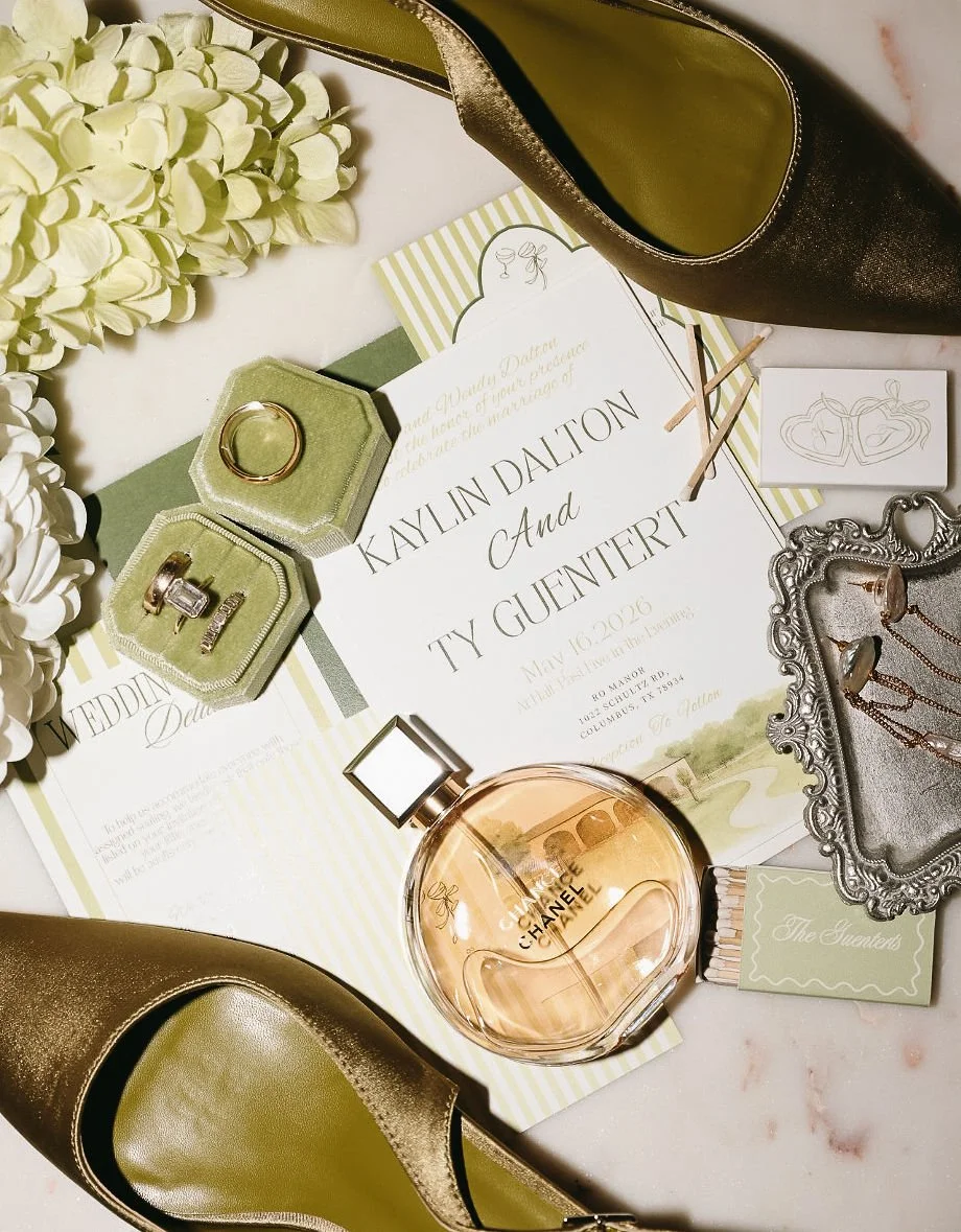 A wedding flatlay with wedding invitation, gold high heel shoes, wedding rings in a green velvet box, a bottle of Chanel perfume, jewelry, a flower bouquet, and decorative ring box on a marble table. 
