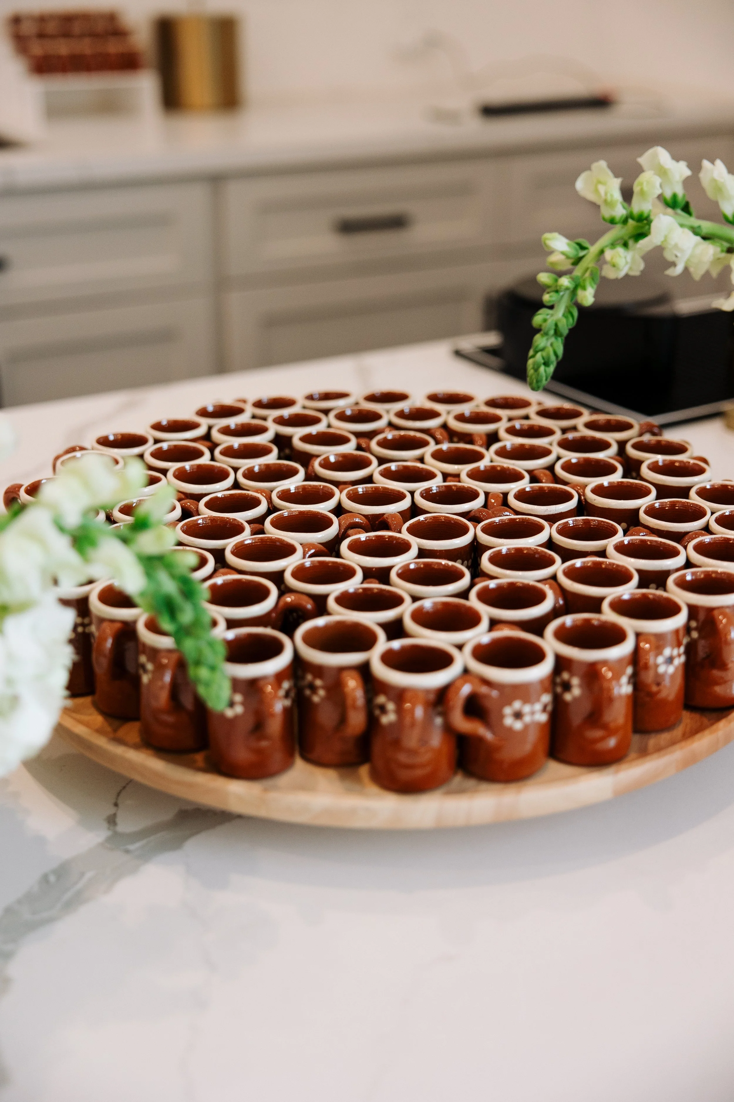 A round wooden tray filled with small ceramic mugs, decorated with white flower patterns, placed on a white marble surface with a blurred background of a wedding bar counter and flowers in the foreground.