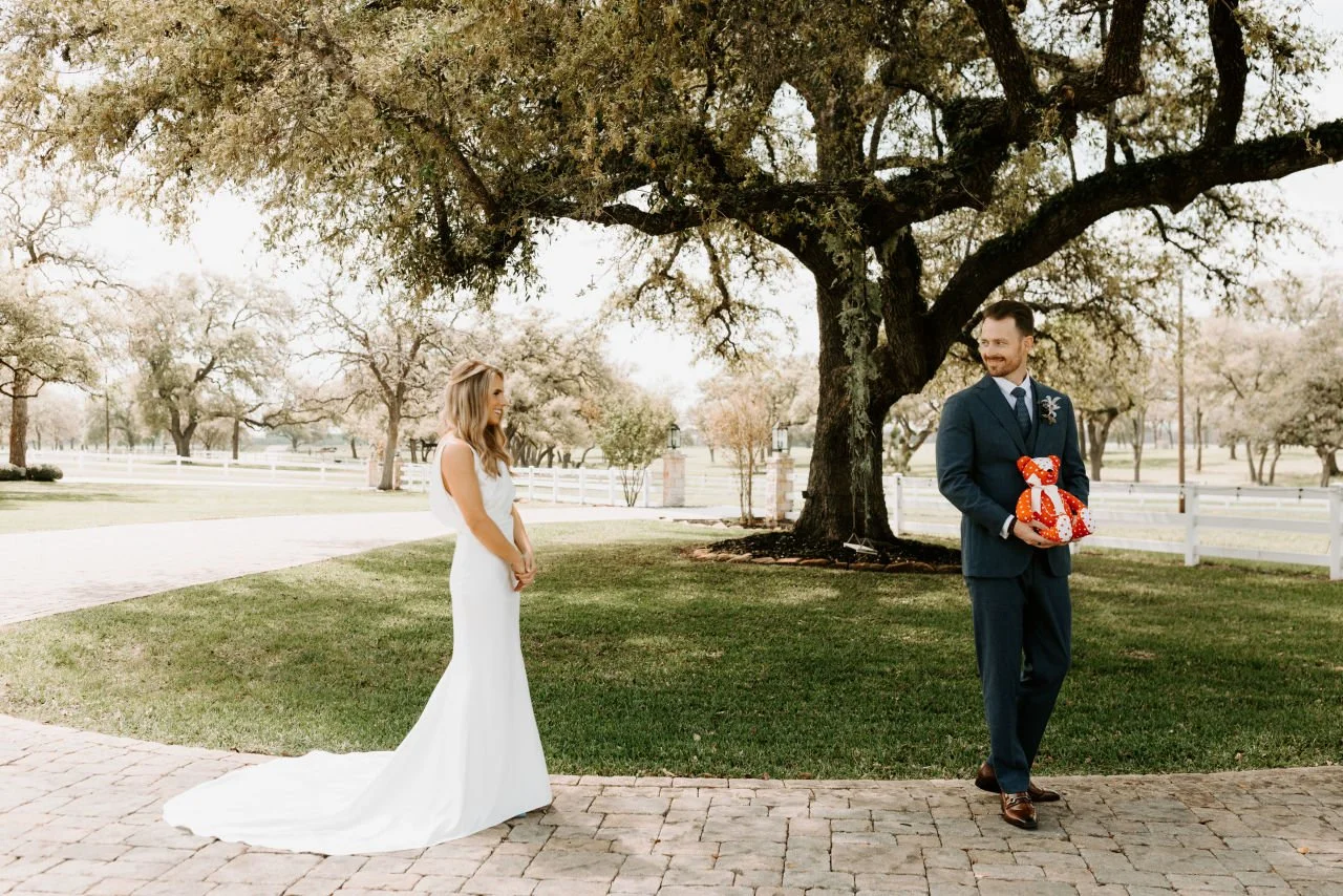A bride and groom outdoors near a large tree. The bride is in a white wedding gown, smiling, while the groom, in a navy suit, holds a red and white polka-dot teddy bear and smiling at her.