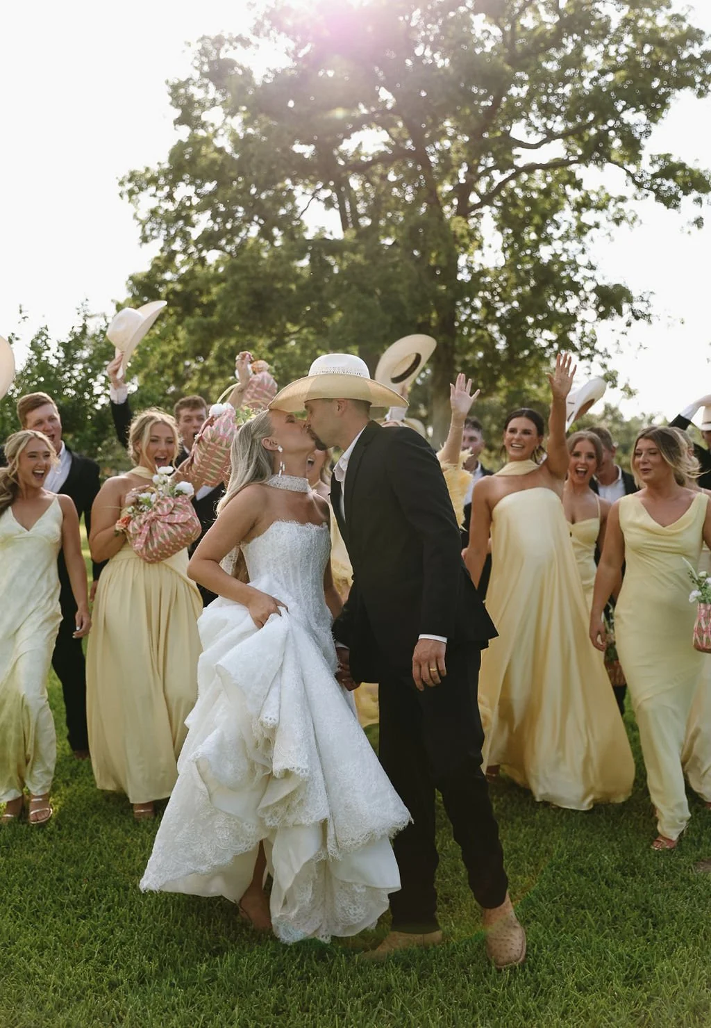 A bride and groom kiss outdoors at their wedding surrounded by bridesmaids and groomsmen. The bridesmaids wear yellow dresses, and the group celebrates with hats in the air, under sunlight and trees.