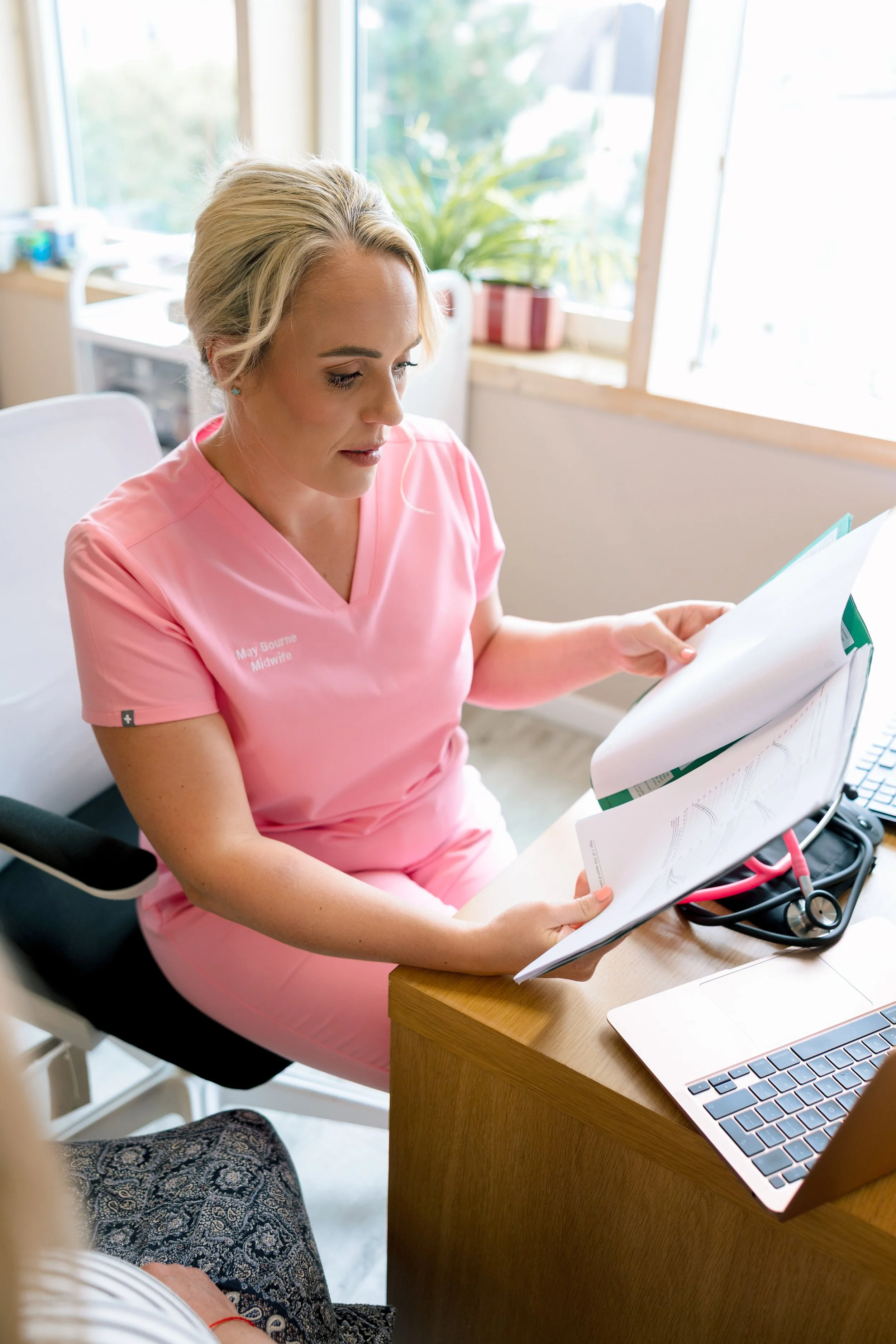 A woman wearing pink scrubs reading a medical chart in an office with a computer on a wooden desk.
