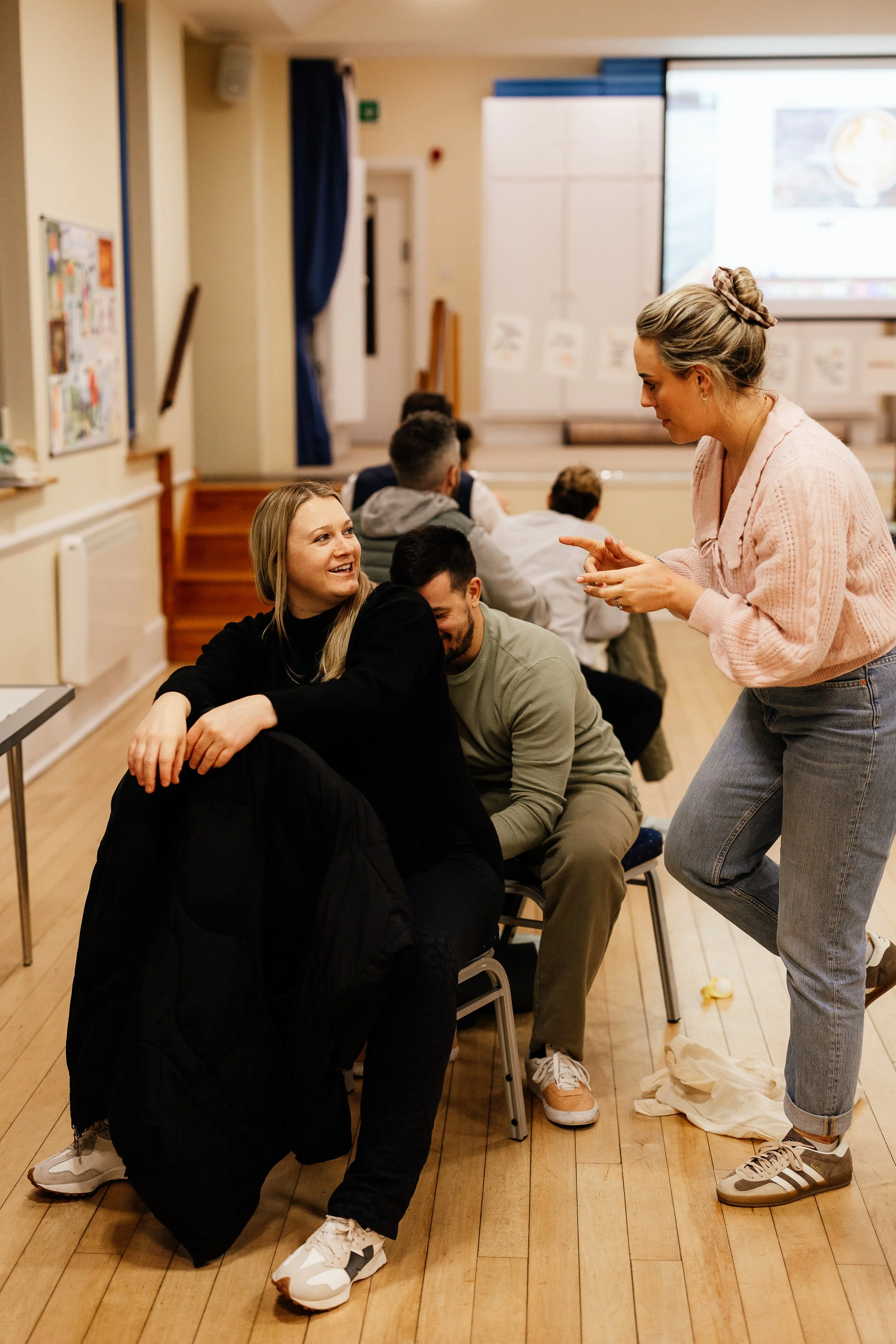 A group of people sitting in a line in a room with a wooden floor, engaging in a conversation. A woman with a bun is standing and talking to a woman with long hair who is smiling. Several others are seated behind them, some facing forward and others turned slightly.