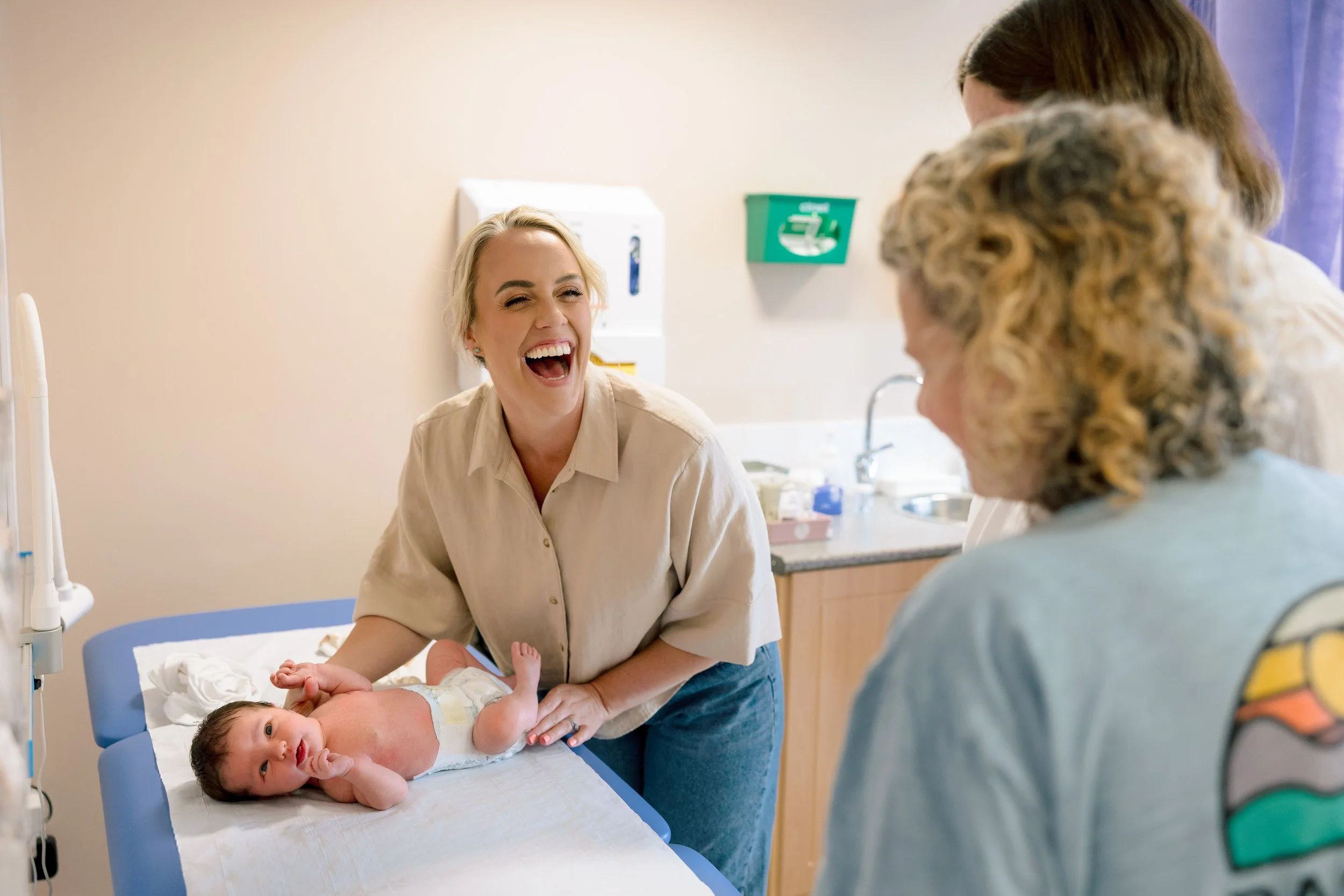 A woman laughing while holding a baby's hand in a hospital room with a nurse or doctor and another adult in the background.