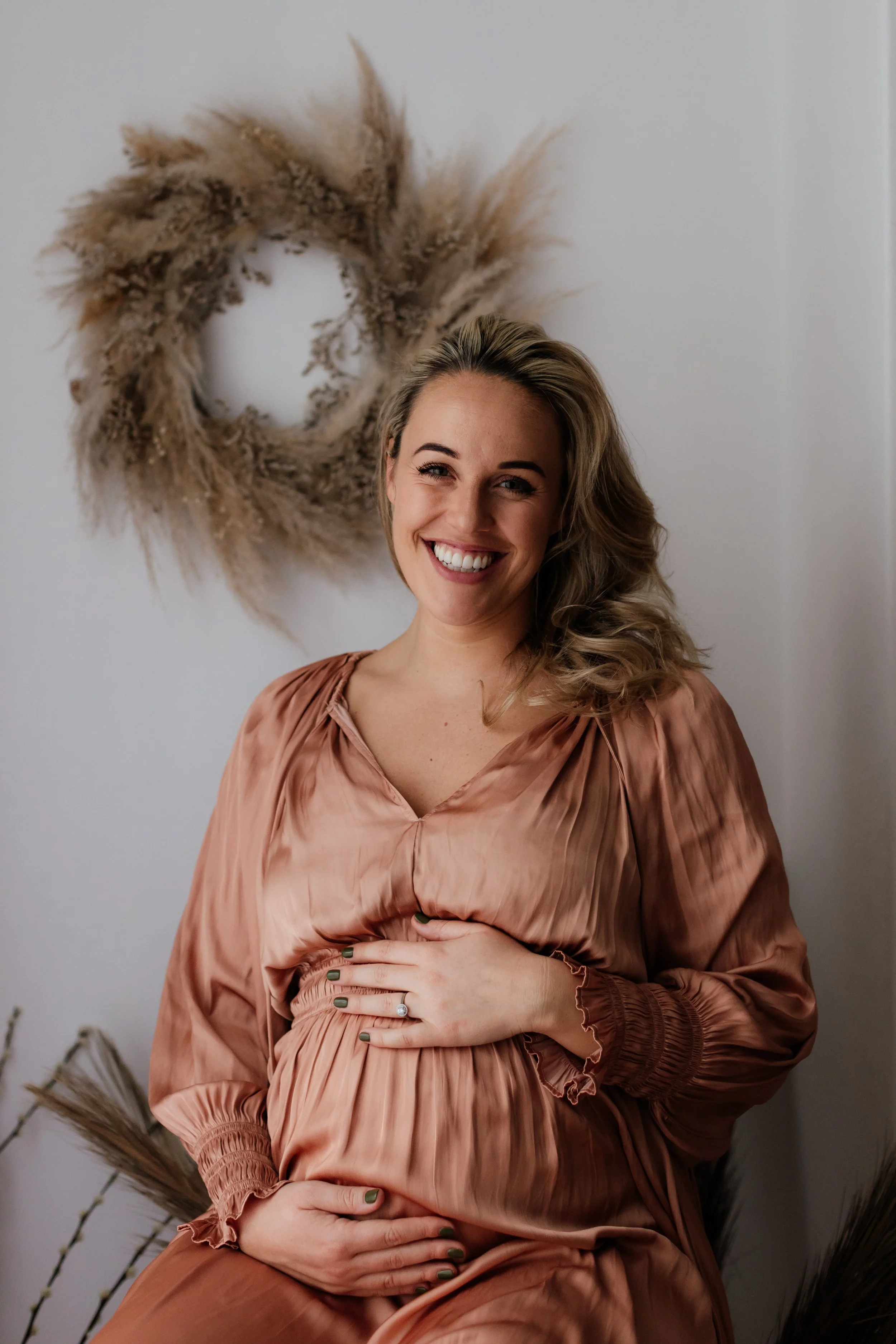 A woman smiling while holding her pregnant belly, standing in front of a decorative wreath made of dried pampas grass and plants on a plain wall.
