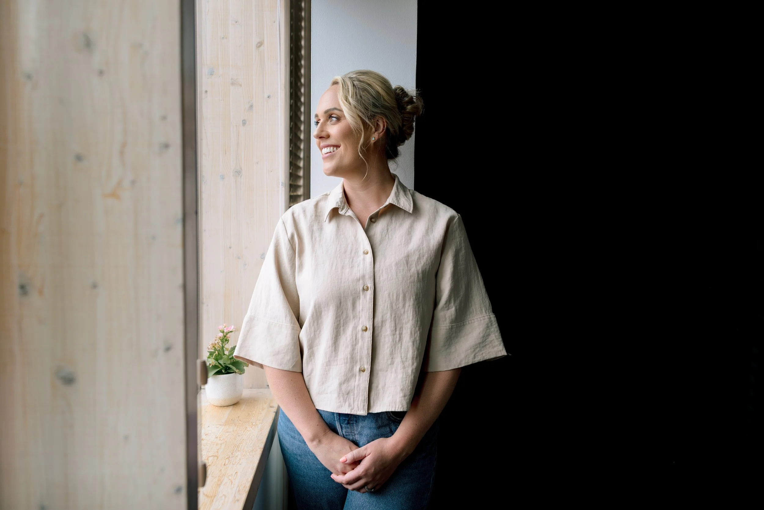 A woman with blonde hair in an updo, wearing a beige button-up shirt and blue jeans, standing by a window with a wooden frame, smiling and looking outside. There is a potted plant on the windowsill.