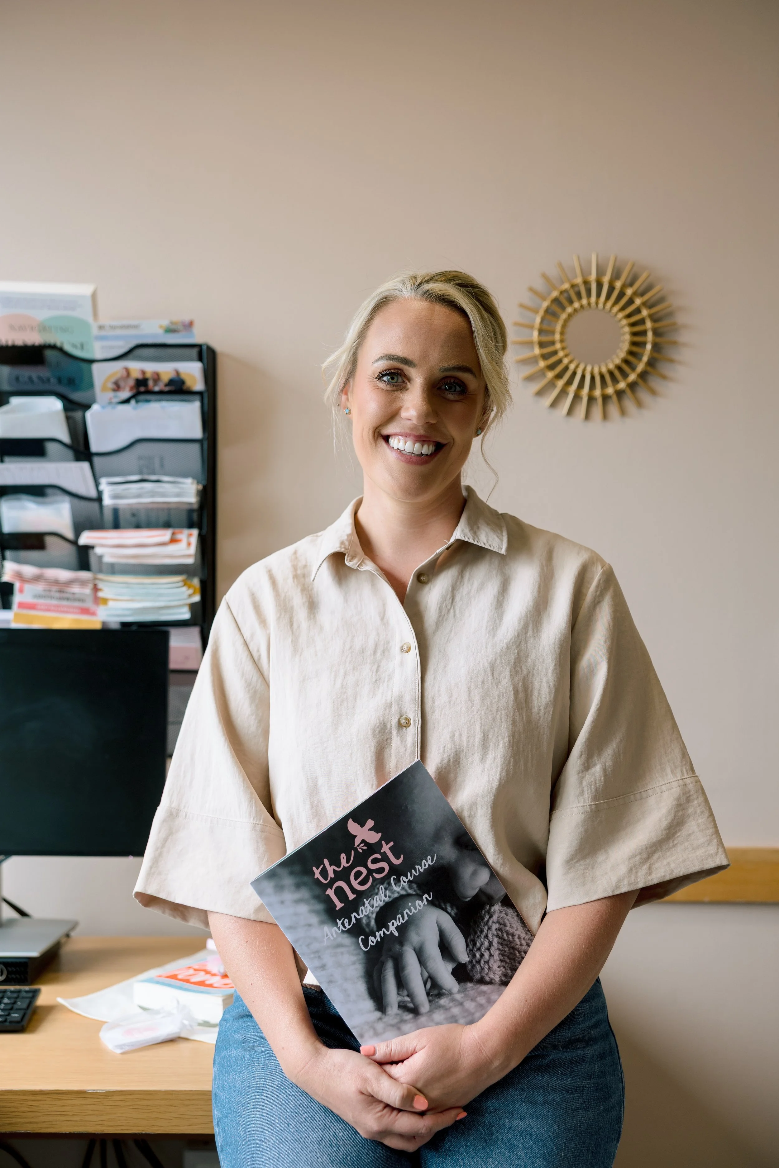 A smiling woman with blonde hair holding a book titled 'the nest' in an office setting with plants, papers, and office supplies in the background.