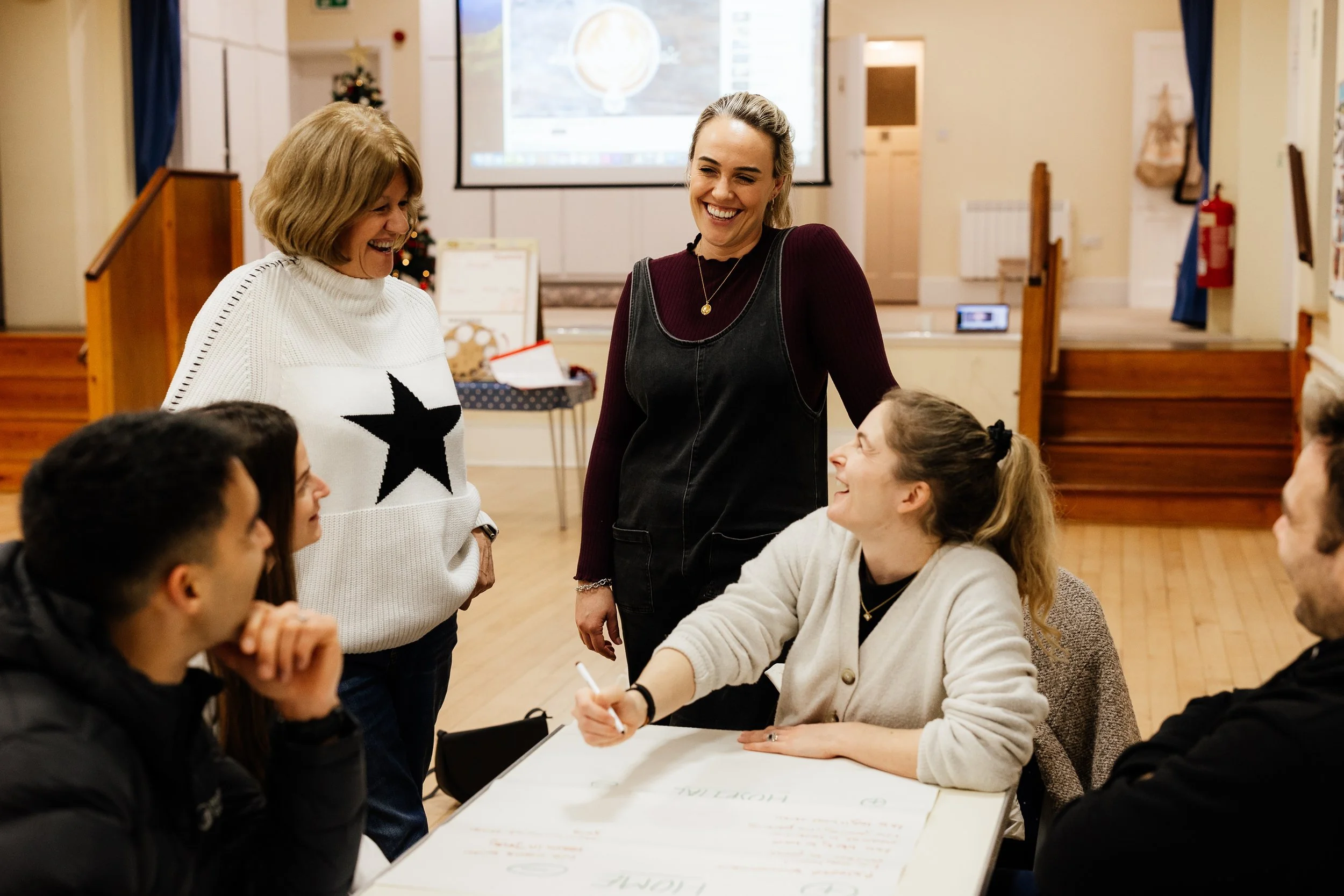 A group of people in a room with a woman standing and smiling, engaging in a discussion with seated individuals around a table that has large sheets of paper with writing on them. There is a decorated Christmas tree and a large projection screen in the background.